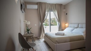 Un dormitorio con una cama, una silla, una mesita con un jarrón de flores y una ventana con cortinas.