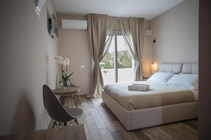 Un dormitorio con una cama, una silla, una mesita con un jarrón de flores y una ventana con cortinas.