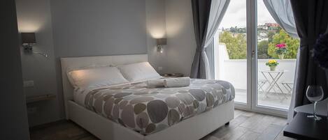 A bedroom with a bed, a bedside table, an air conditioning unit, a window with curtains, and a balcony with a table and chairs.