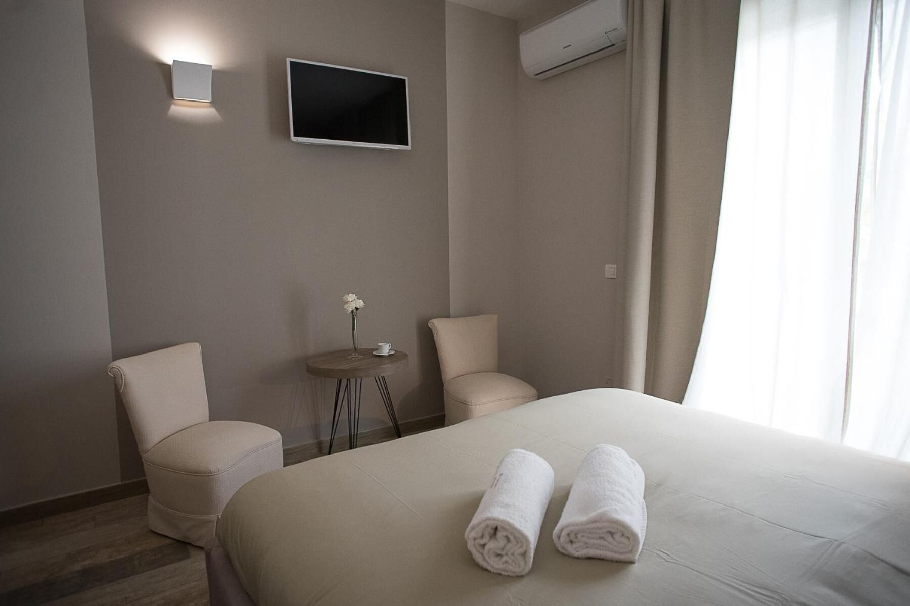A hotel room with a bed, two chairs, a small table with a vase, a TV mounted on the wall, and an air conditioning unit.