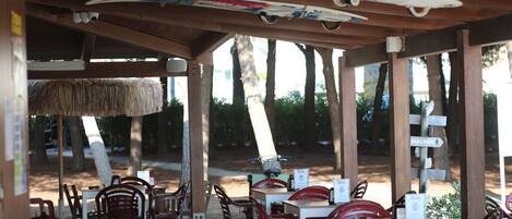 An outdoor dining area with wooden tables and chairs, a thatched roof, and surfboards hanging above.