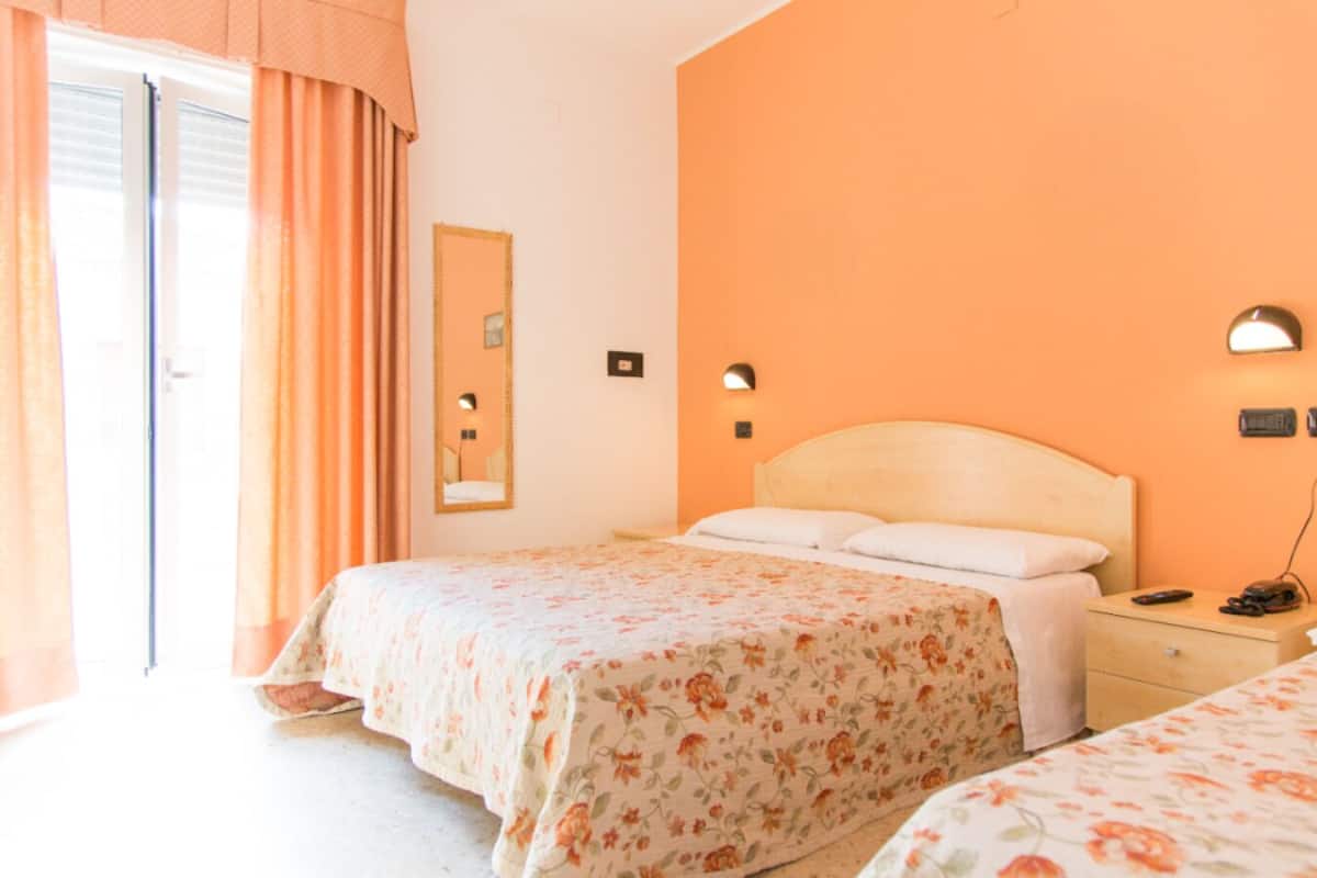 A hotel room with two beds, a large window with curtains, a wall-mounted mirror, and a bedside table with a lamp.