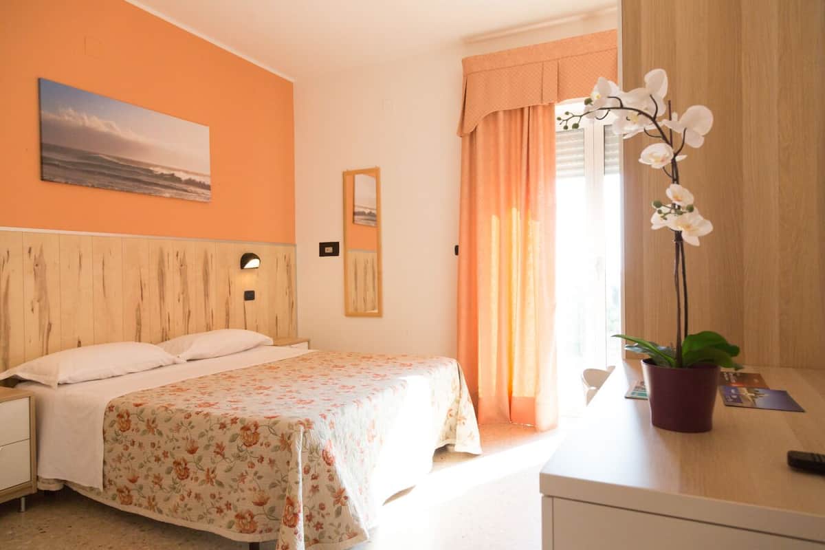 A hotel room with a wooden headboard, a bed with floral bedding, a nightstand, a desk with a potted orchid, and a window with curtains.