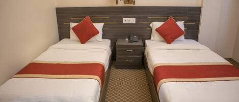 A hotel room with two single beds, each with a red pillow and a white blanket. There is a nightstand with a phone and a lamp between the beds. The room has a patterned carpet and a wall-mounted light fixture above the beds.