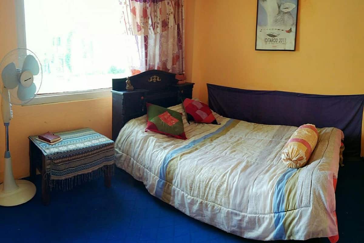 A bedroom with a bed, a bedside table, a standing fan, a window with curtains, and a poster on the wall.
