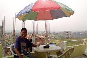 Un hombre sentado en una mesa con un paraguas colorido, con vistas a una obra en construcción y edificios al fondo.