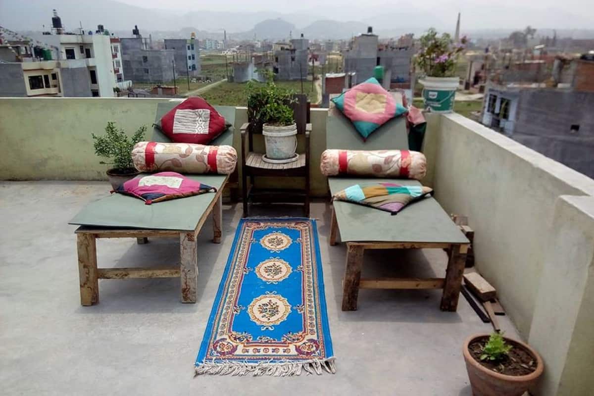 A rooftop terrace with two wooden tables, colorful cushions, a blue patterned rug, and potted plants.