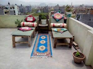 Una terraza en la azotea con dos mesas de madera, cojines de colores, un tapete azul estampado y plantas en macetas.
