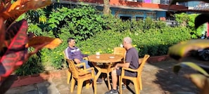 Two people seated at a round wooden table outdoors with a garden in the background.