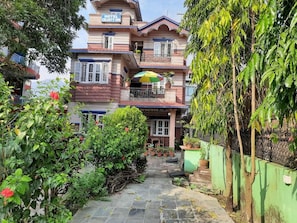 A hotel with a multi-story structure, a balcony with a colorful umbrella, and a garden area with potted plants.
