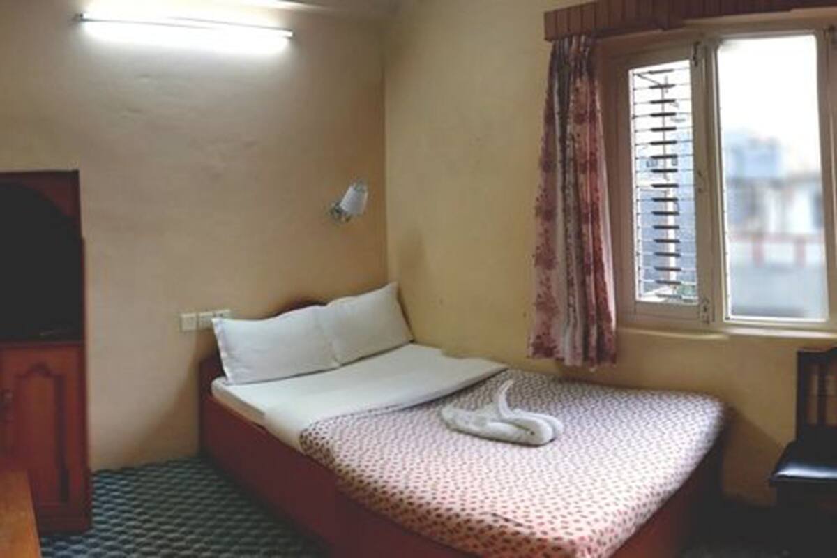 A small, single-bed room with a patterned bedspread, a window with curtains, and a wall-mounted light fixture.