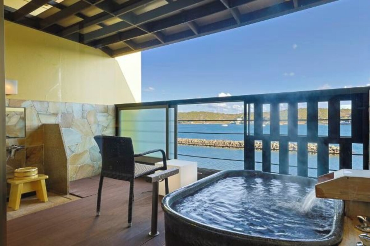 A hot tub with a wooden bench and a small stool, a stone wall, and a view of the sea through a balcony railing.