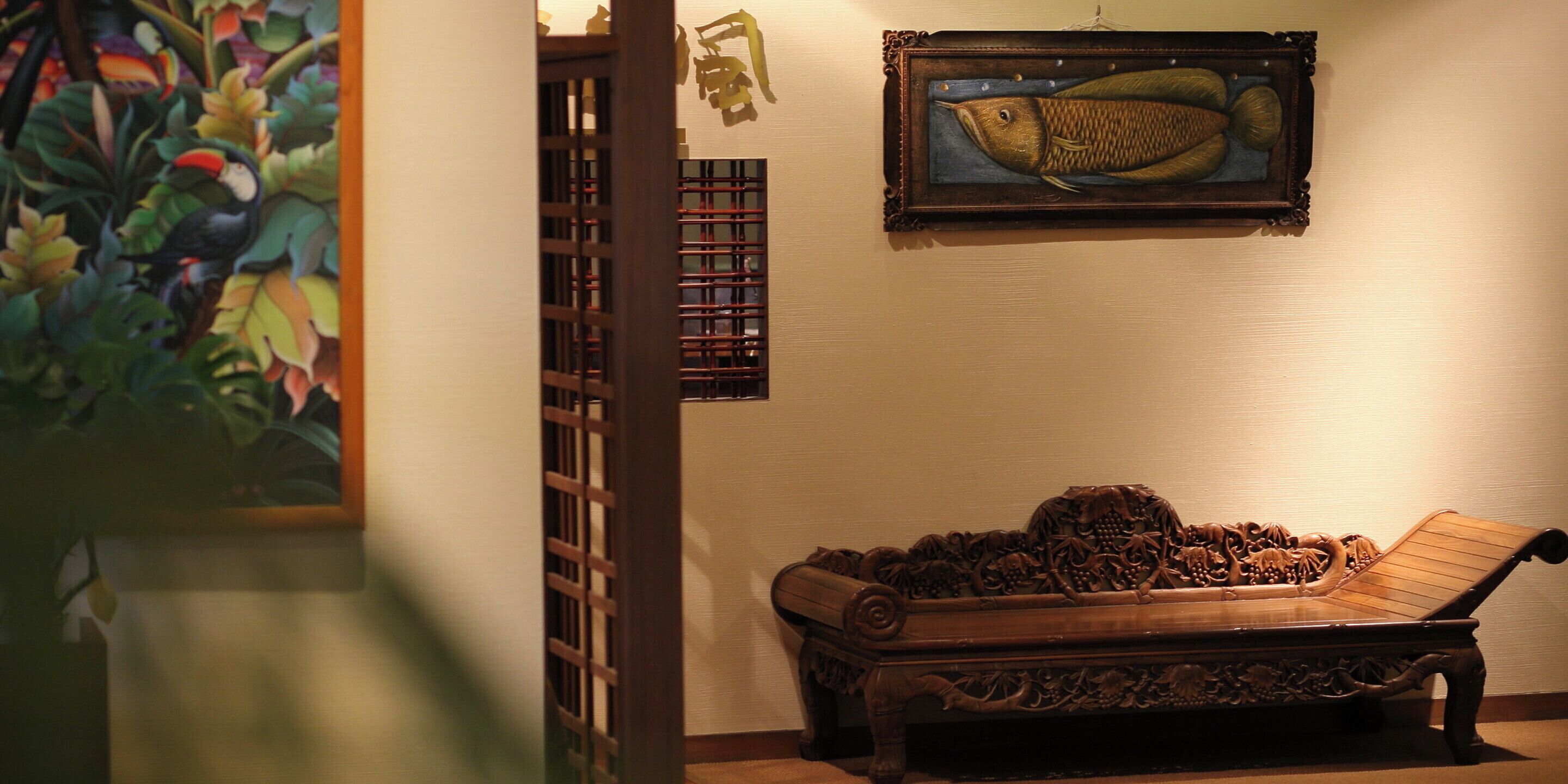 Un banc en bois traditionnel orné de sculptures complexes, un tableau encadré représentant un poisson et une porte en treillis de bois.