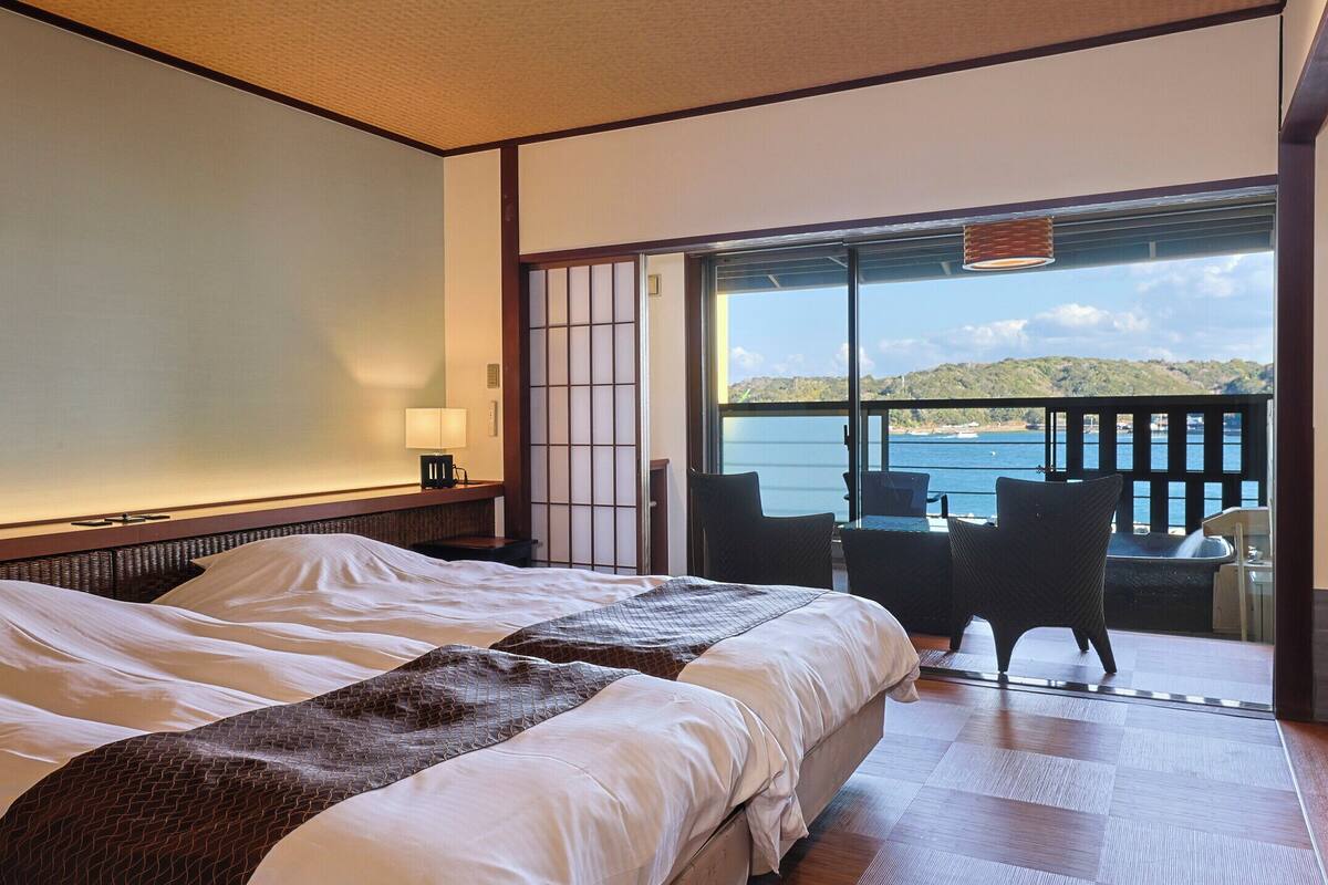 A hotel room with two beds, a view of the sea, and a balcony with chairs.