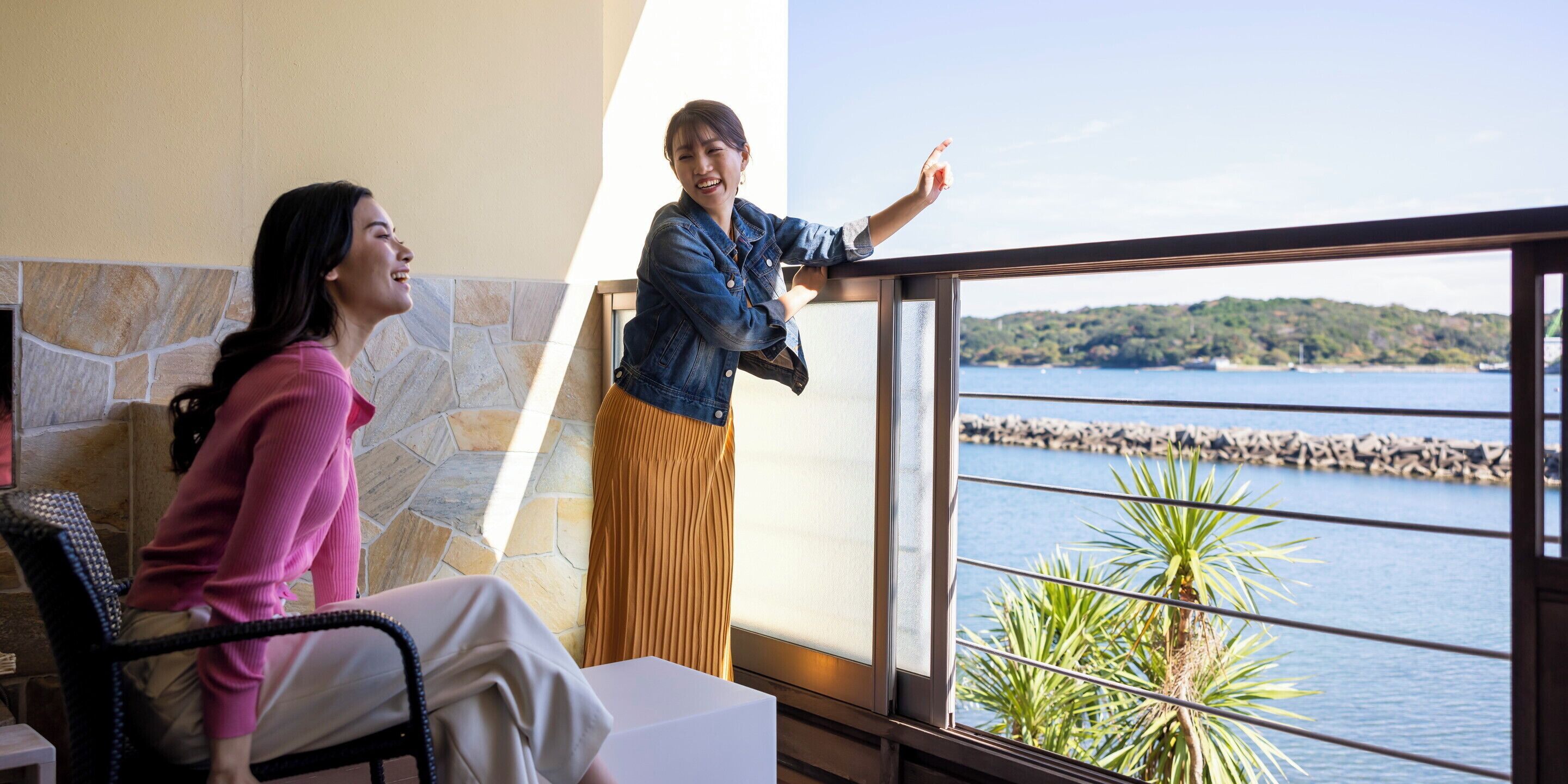 A room with a stone wall, a large window with a view of a body of water and a distant shoreline, and a person standing by the window.