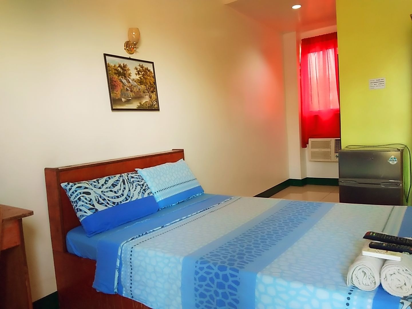 A hotel room with a bed, a painting on the wall, a red curtain, and a refrigerator.
