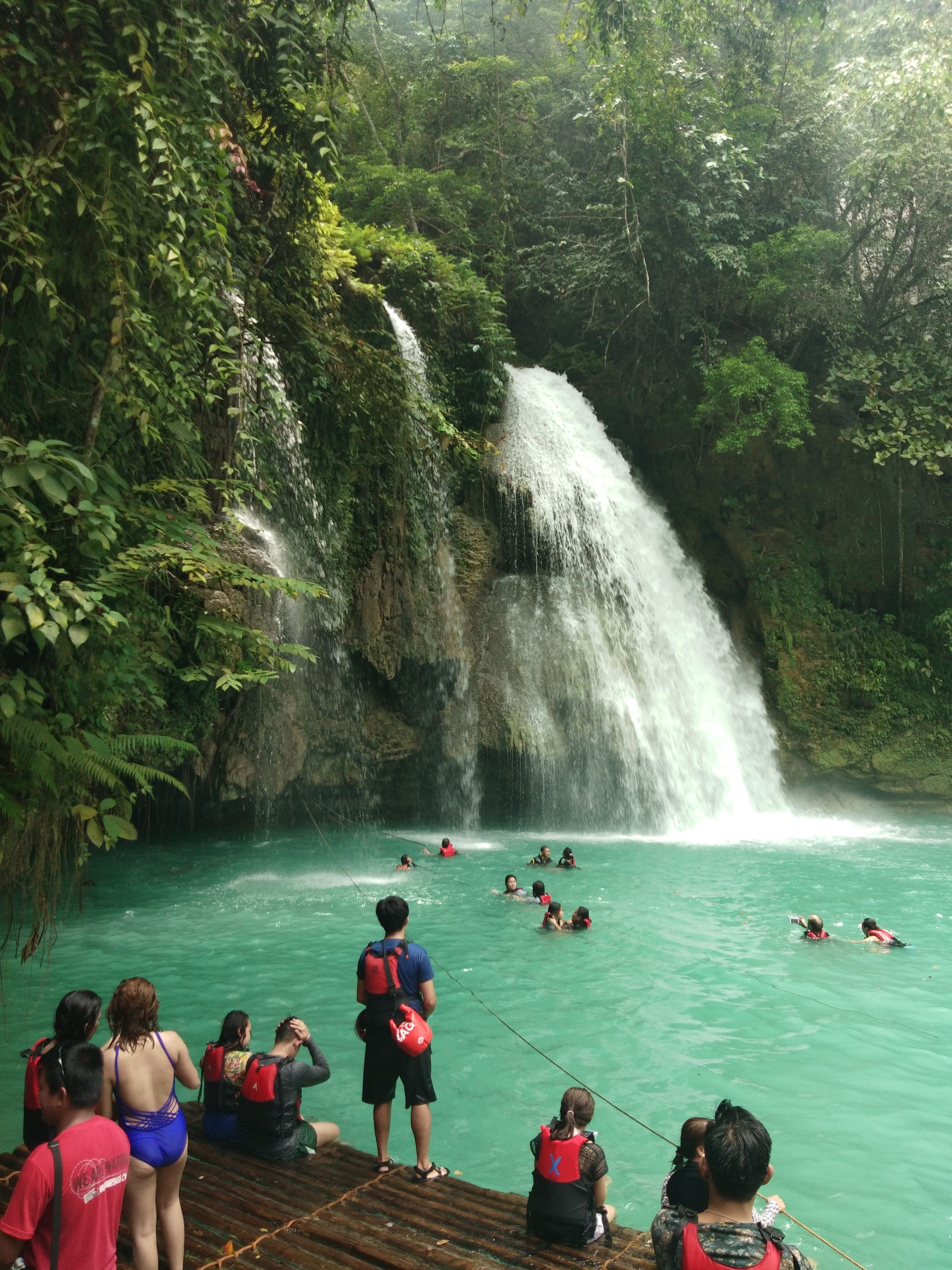 Una cascada en un entorno de bosque exuberante y verde, con gente nadando y parada sobre una plataforma de madera.