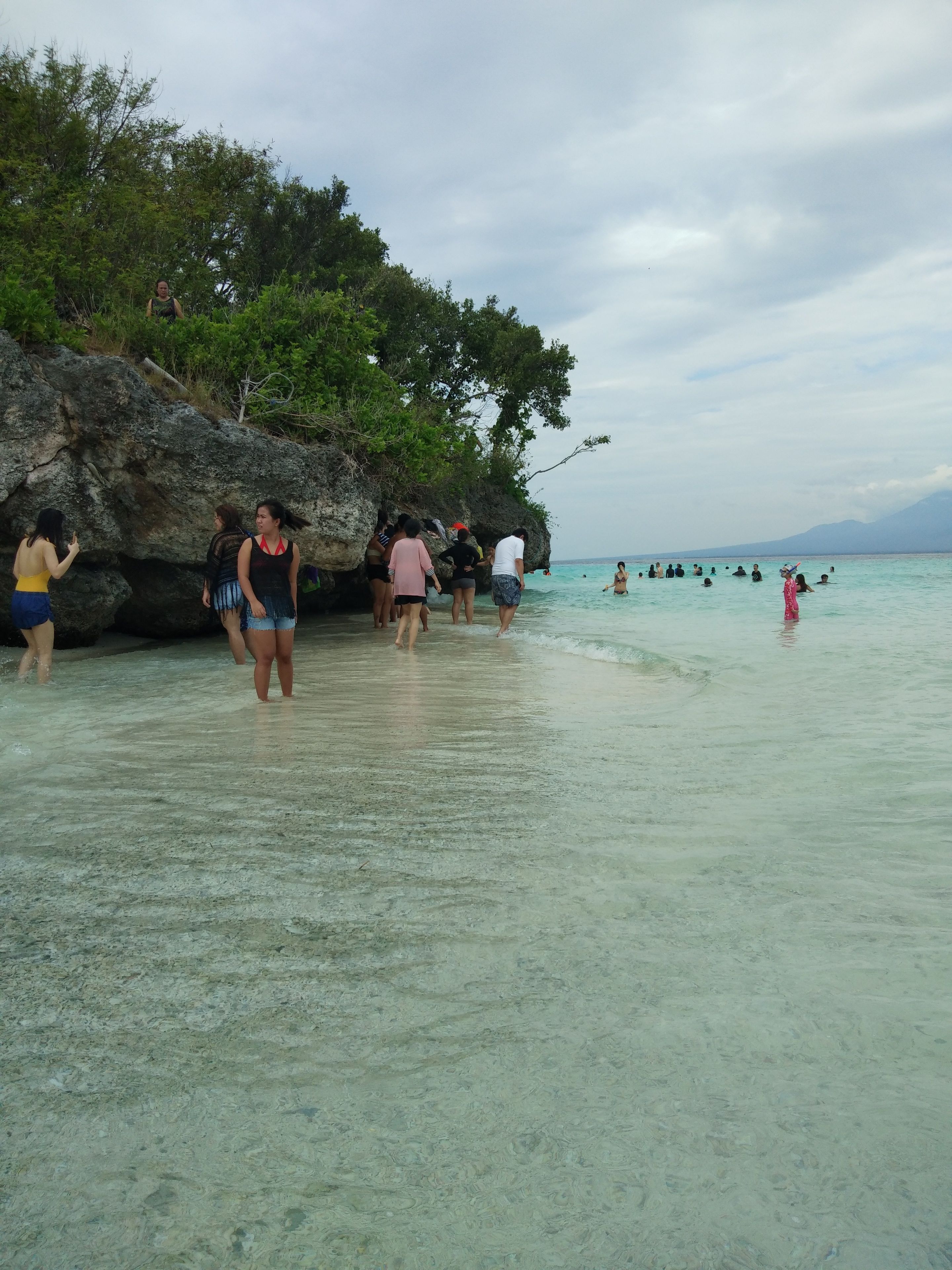 A beach with clear water, rocky cliffs, and people swimming and walking along the shore.