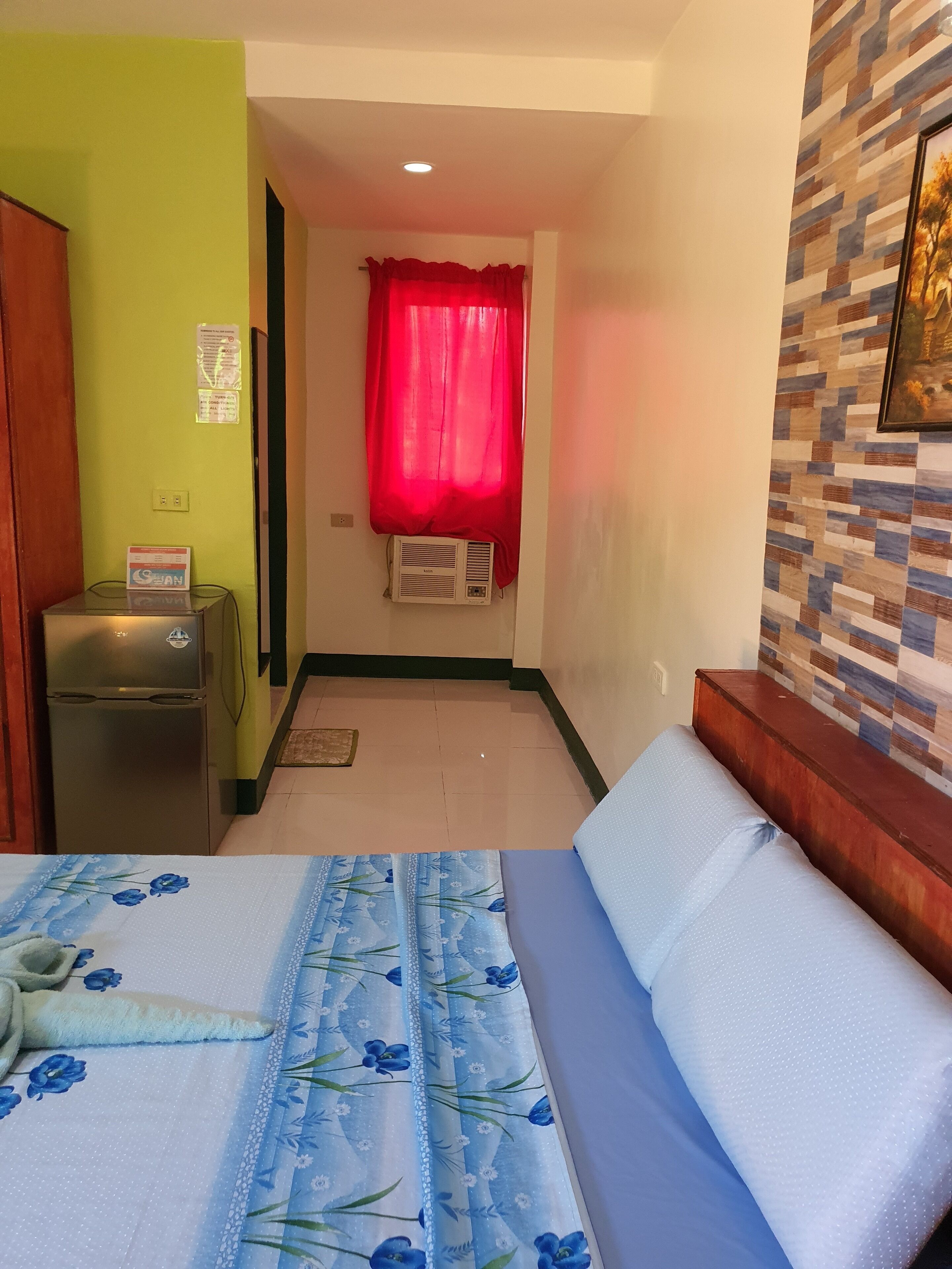Habitación de hotel con una cama, un refrigerador pequeño, aire acondicionado instalado en la pared, una cortina roja y una pared de azulejos con un cuadro.