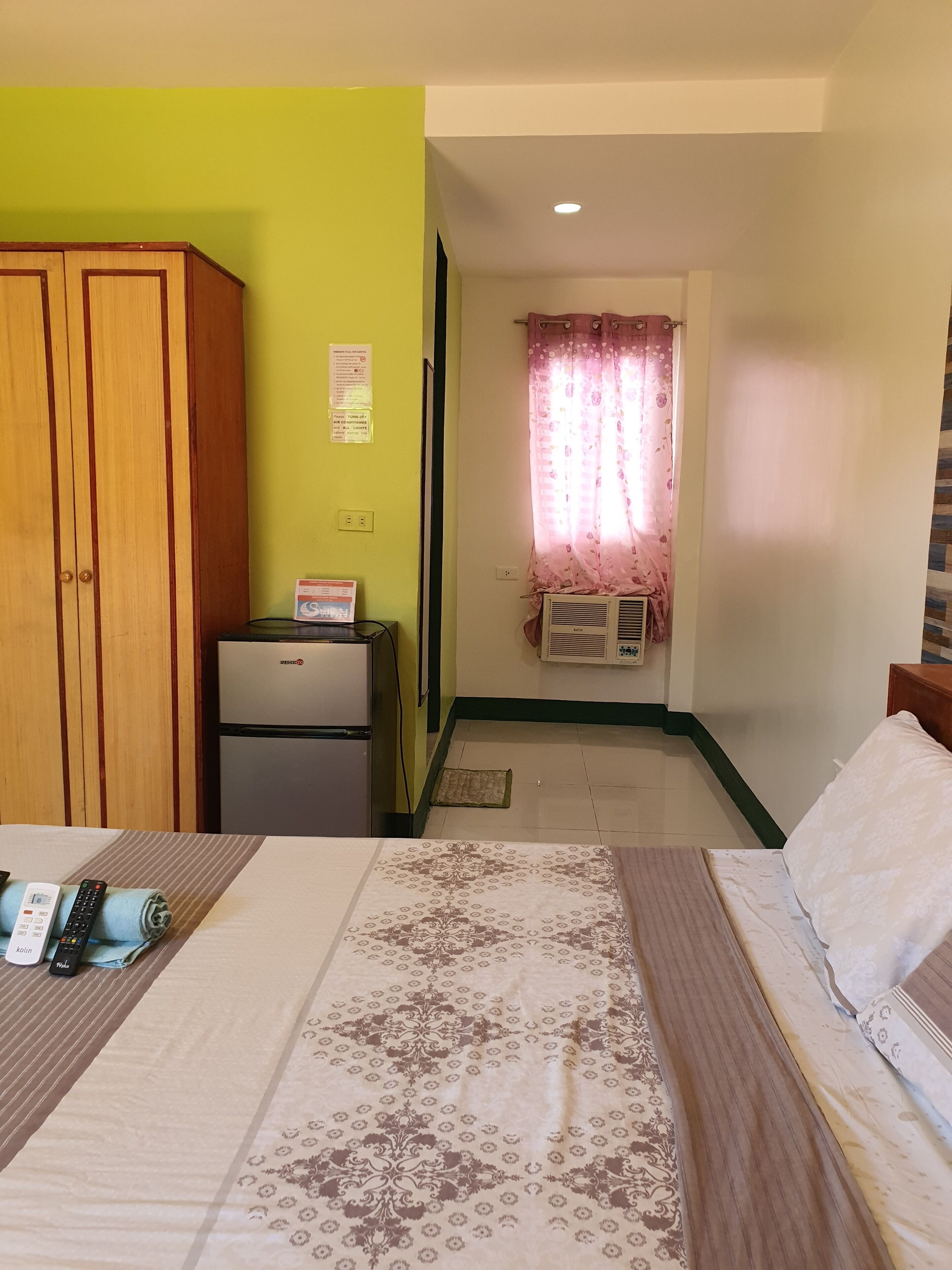A hotel room with a bed, a wardrobe, a small refrigerator, and a window with curtains.