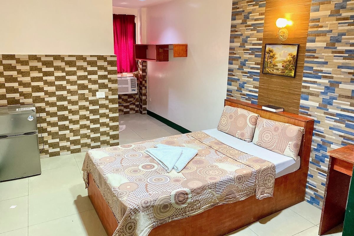 A hotel room with a bed, a desk, a refrigerator, and a patterned wall.