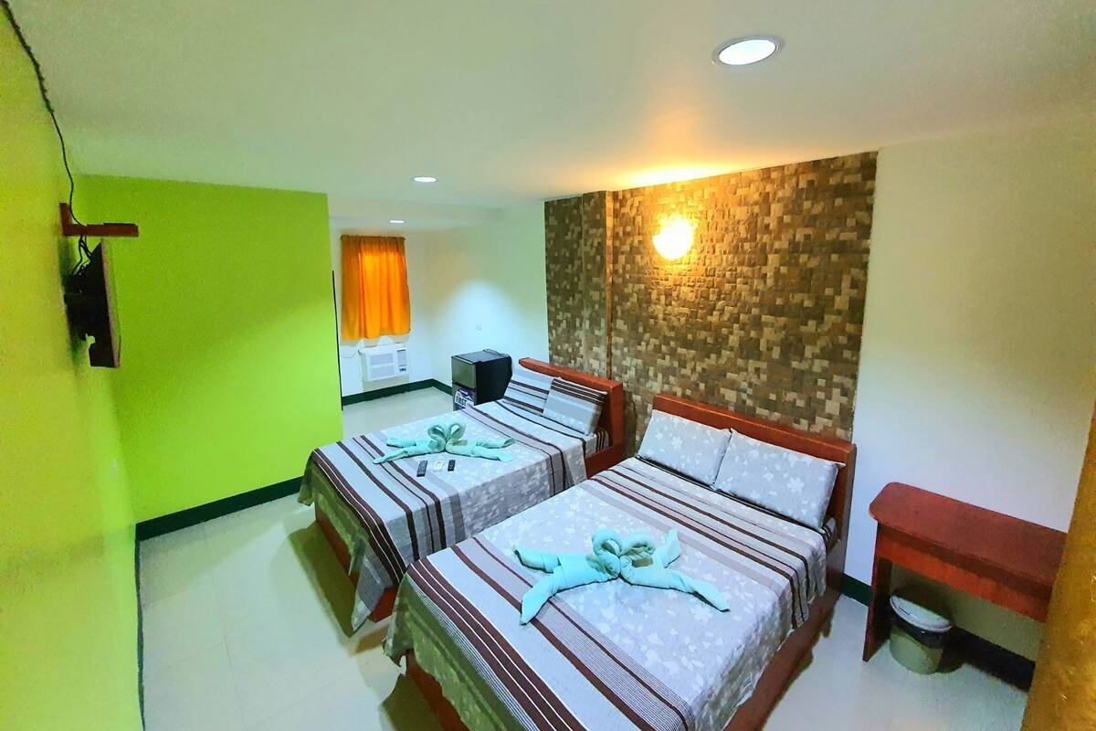 A hotel room with two beds, a small desk, a wall-mounted TV, and a wall light.