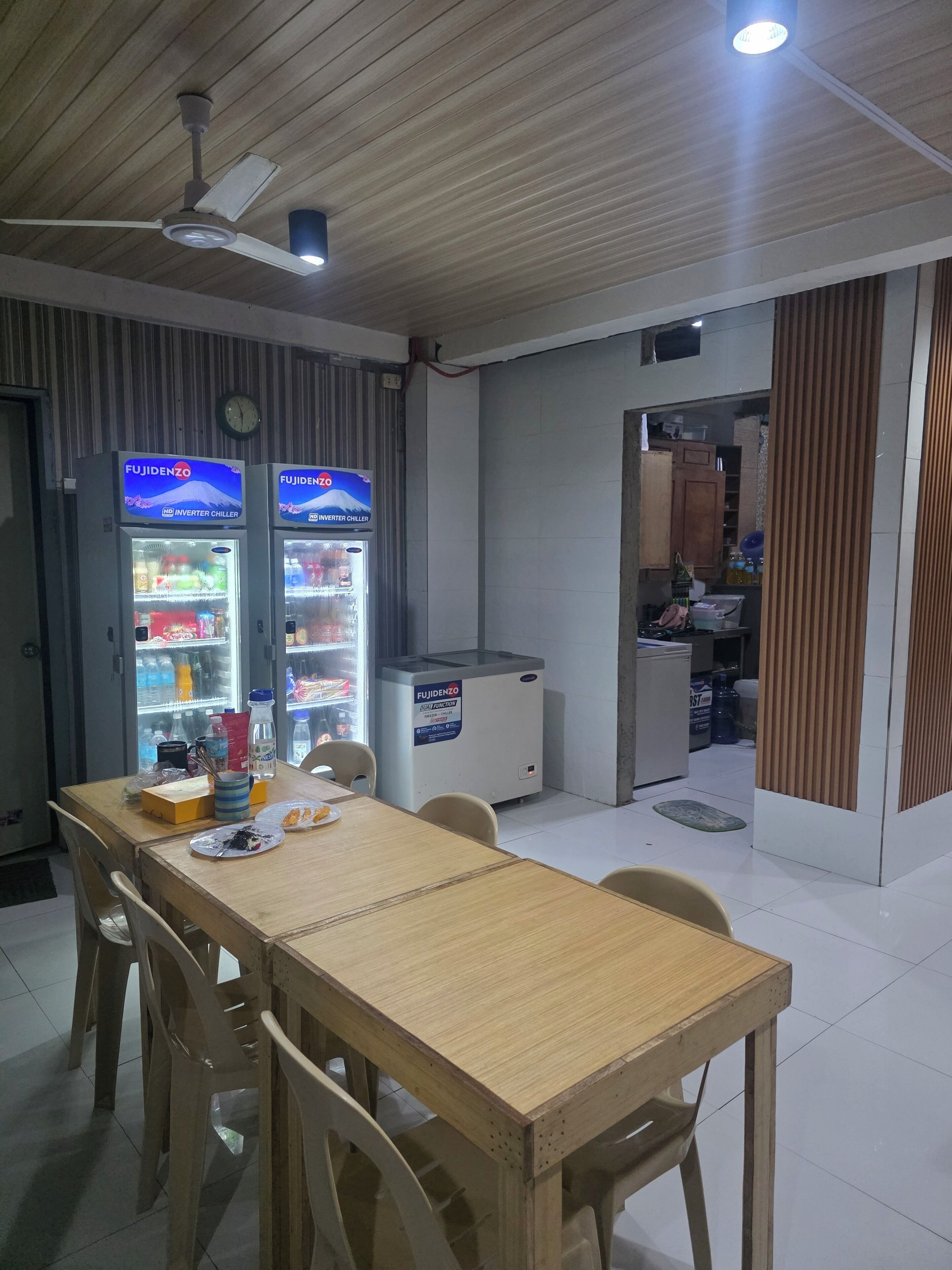 Un coin repas avec une table et des chaises en bois, deux réfrigérateurs de la marque Fujidenzo, un ventilateur de plafond et une horloge murale.