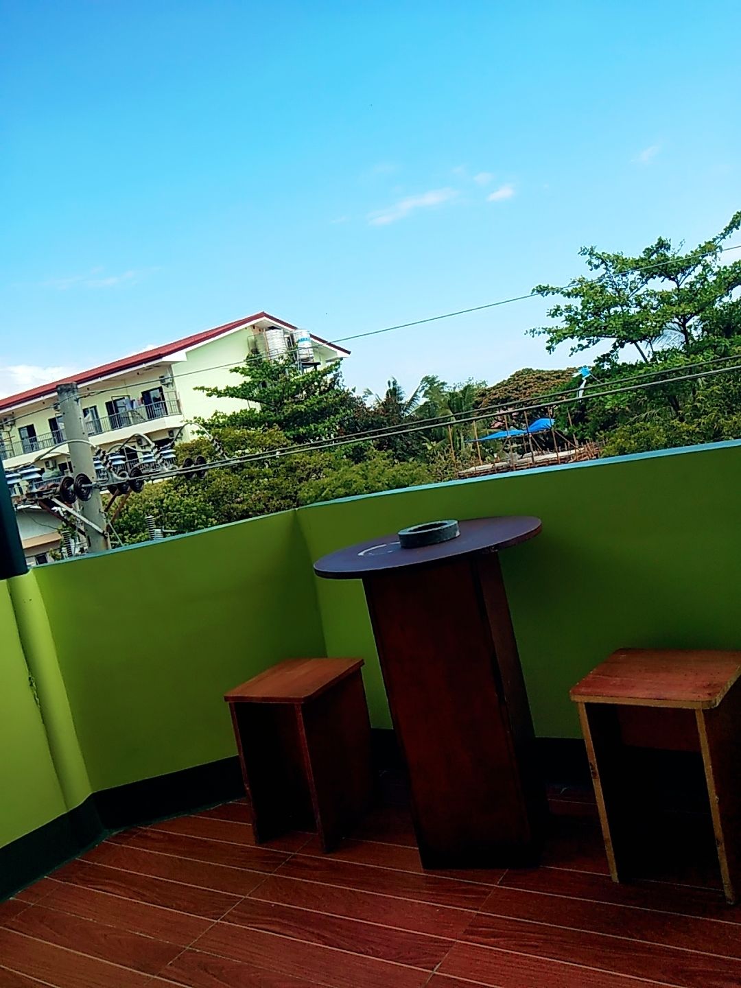 A balcony with a table and two chairs, a green wall, and a view of a building with a red roof.