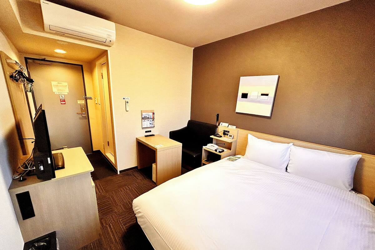 A hotel room with a bed, a desk, a chair, a mirror, and an air conditioning unit.