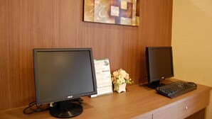 Two computer monitors on a wooden desk with a keyboard and a small vase of flowers. - Hotel Route Inn Anan (Anan)