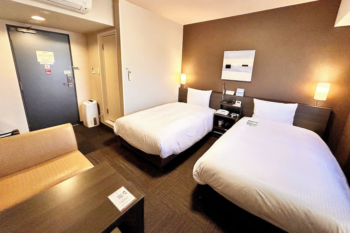 A hotel room with two beds, a desk, a chair, a bedside table, a lamp, and a wall-mounted TV.