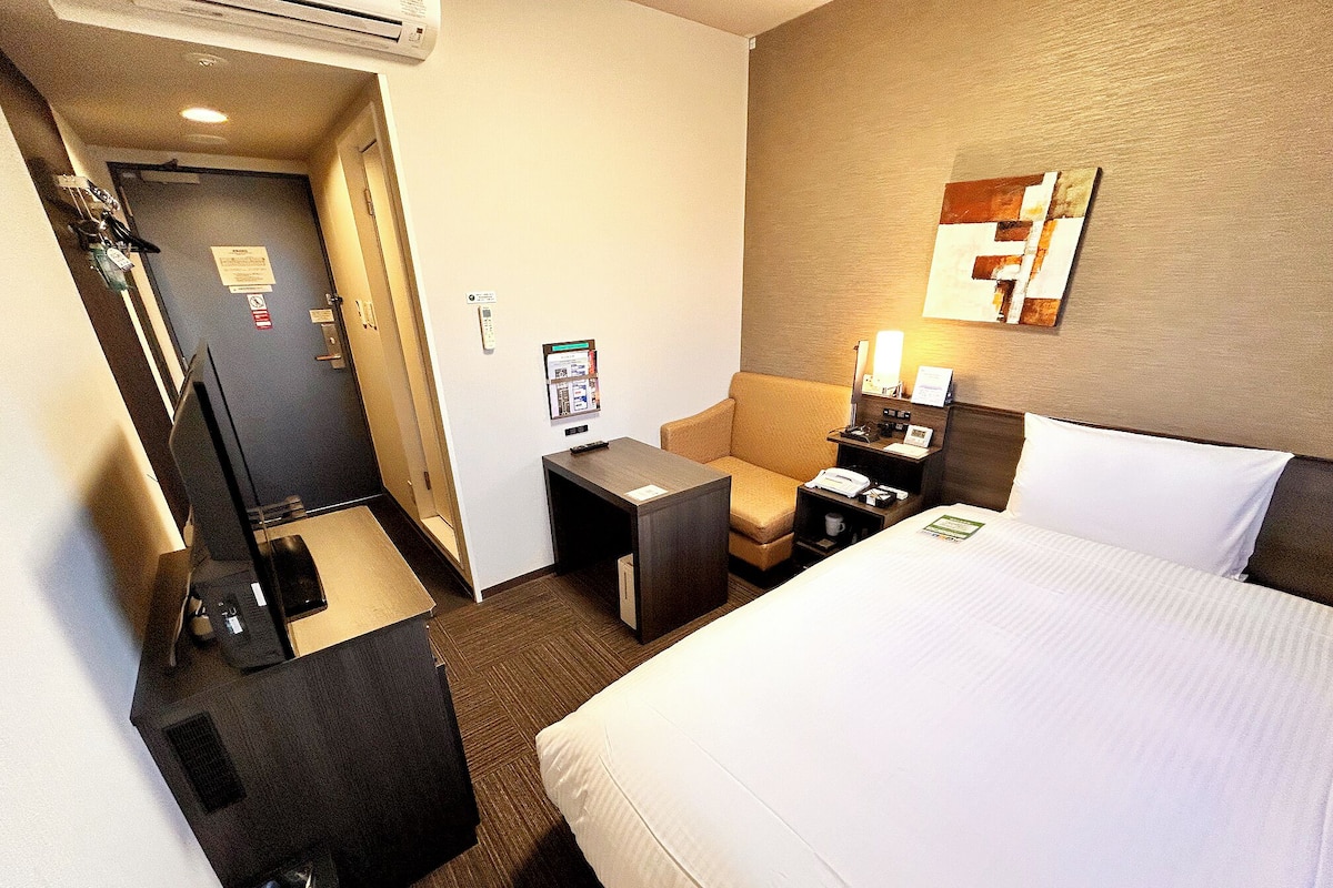 A hotel room with a bed, a desk, a chair, a small table, and a mirror on the wall.
