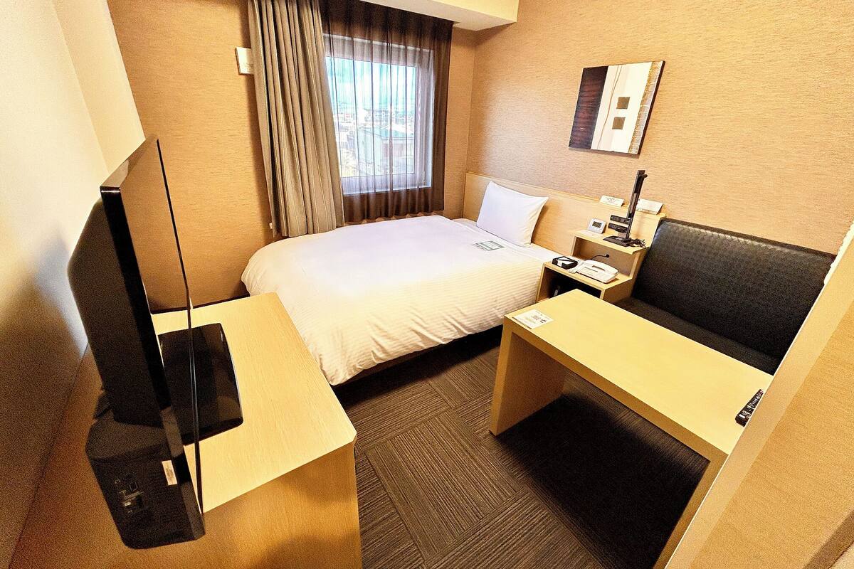 A hotel room with a bed, bedside table, desk, chair, lamp, and a window with curtains.