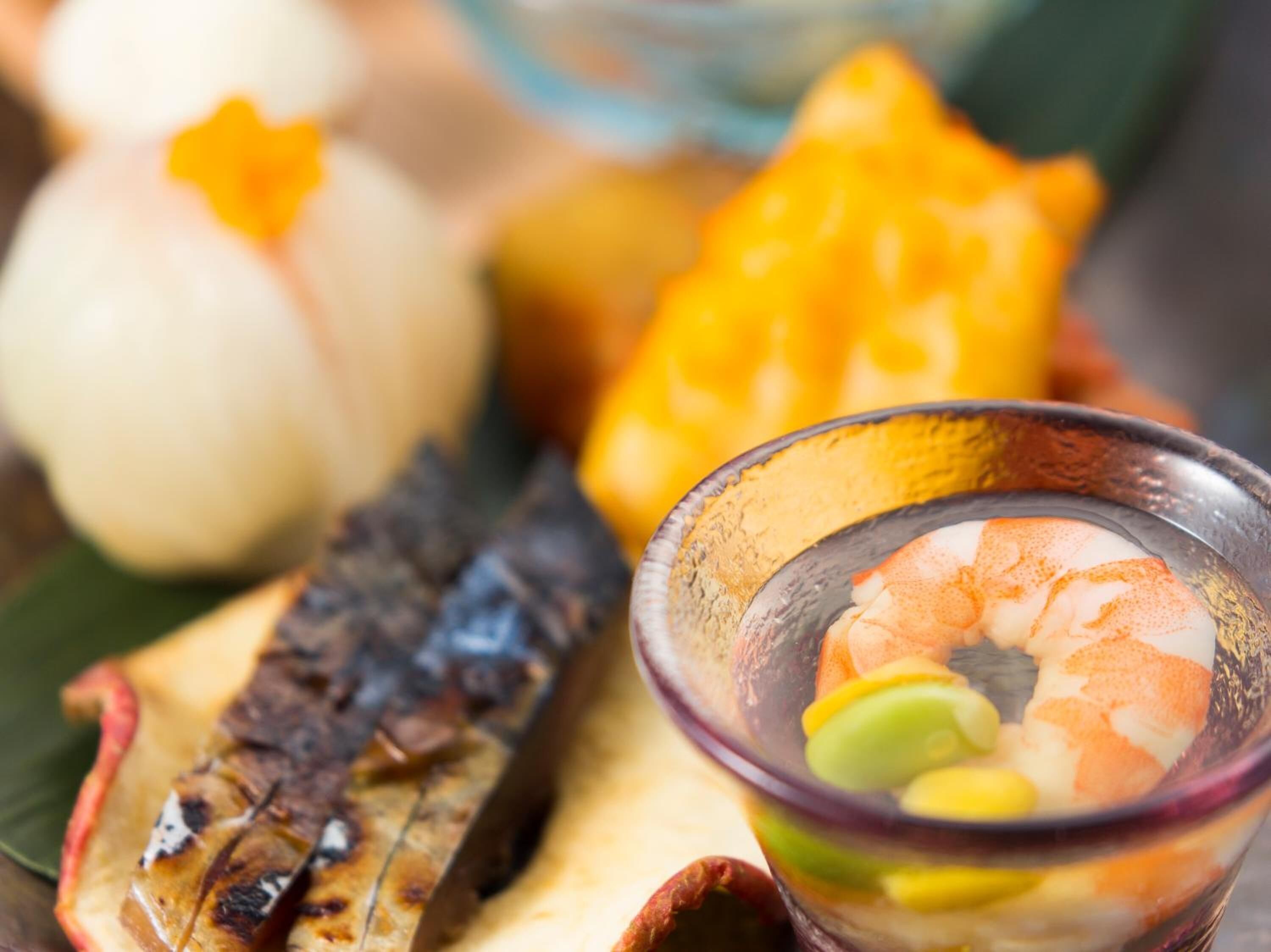 A bowl of shrimp with green and yellow garnish, a grilled fish, and a white and orange dish.