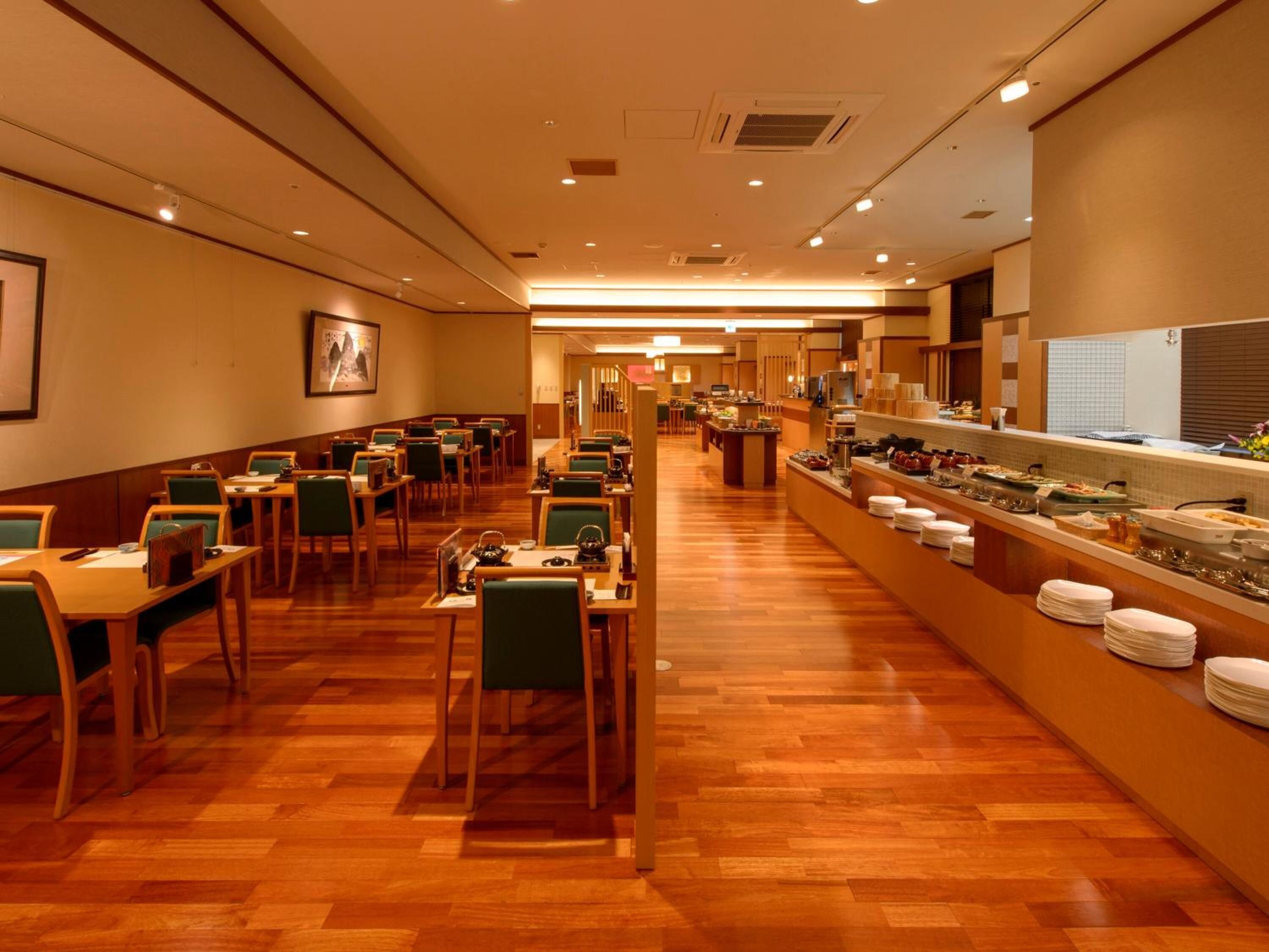 A hotel dining area with wooden flooring, green chairs, and a buffet counter with various food items.