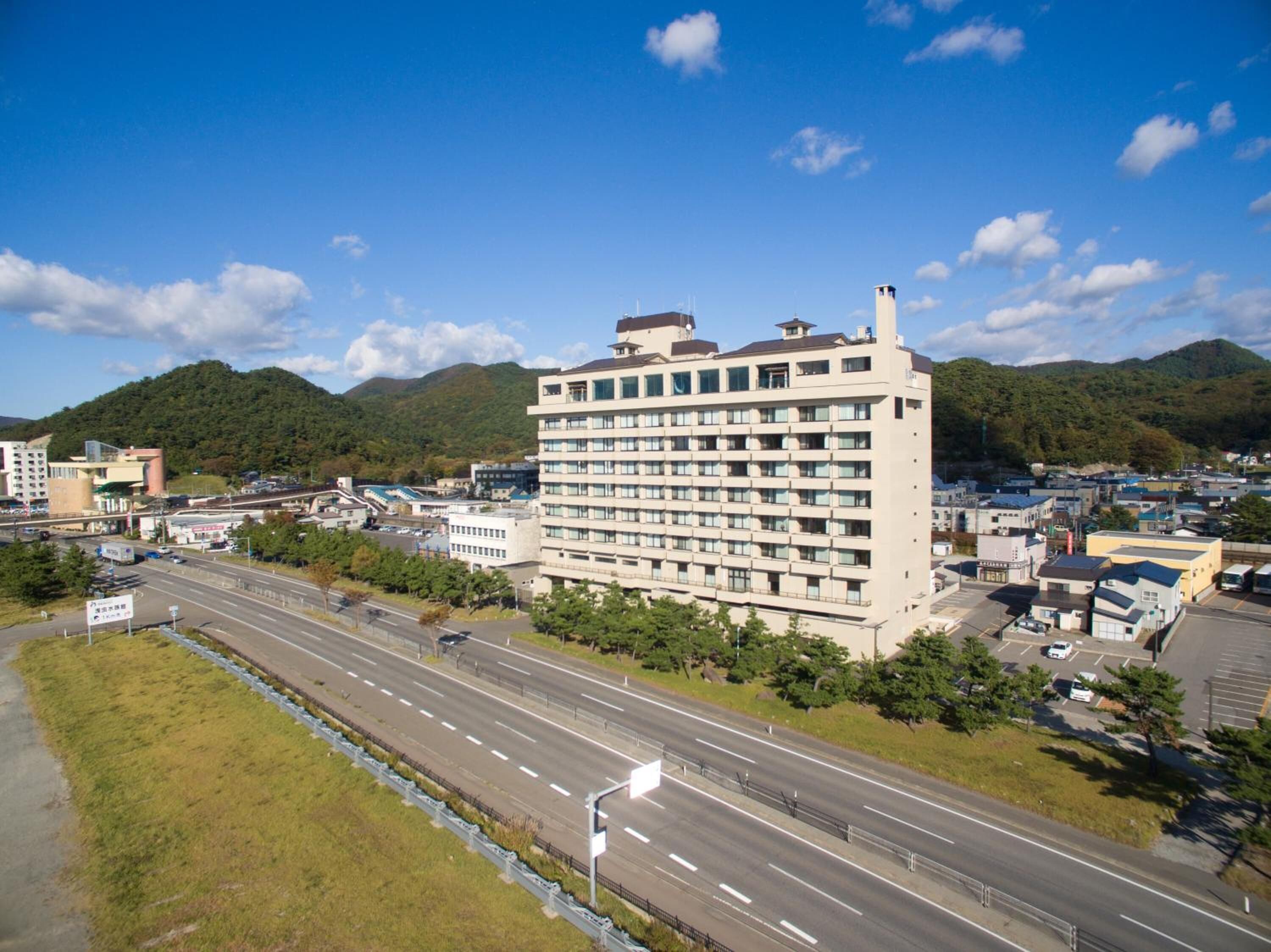 A multi-story hotel with a modern design, located near a highway and surrounded by greenery and mountains.