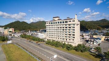 A multi-story hotel with a modern design, located near a highway and surrounded by greenery and mountains.