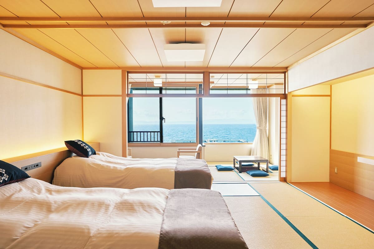 Hotellihuone, jossa on kaksi sänkyä, suuret ikkunat, joista avautuu näkymä merelle, tatamimatto ja perinteinen japanilainen liukuovi.