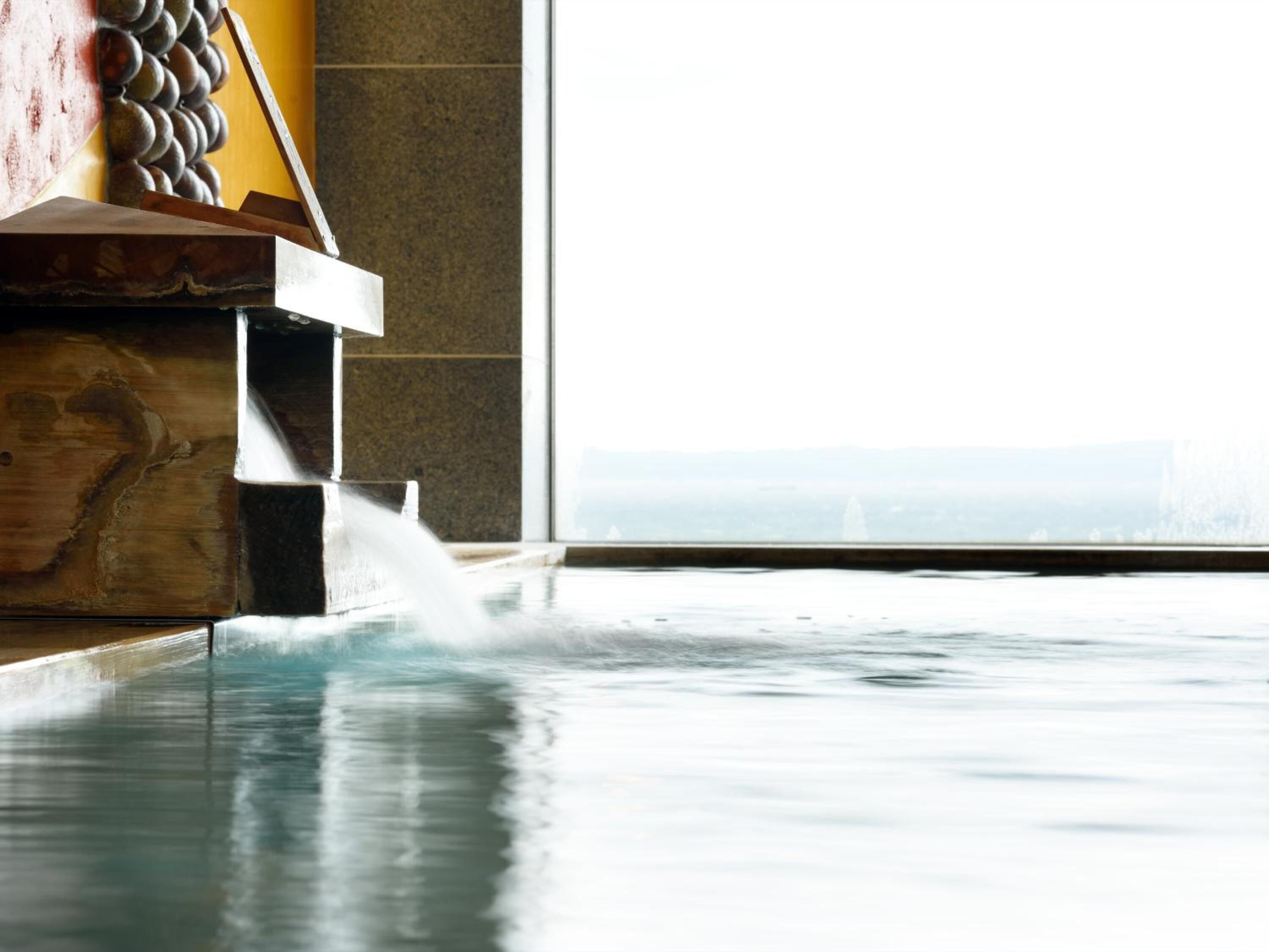 A spa area with a water feature, a wooden bench, and a view of a cityscape through a large window.