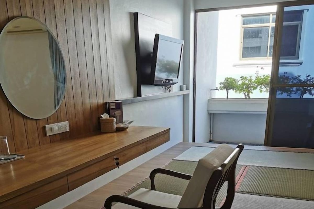 Een moderne woonkamer met een houten bankje, een flatscreen televisie, een ronde spiegel en uitzicht op de omgeving door een groot raam.