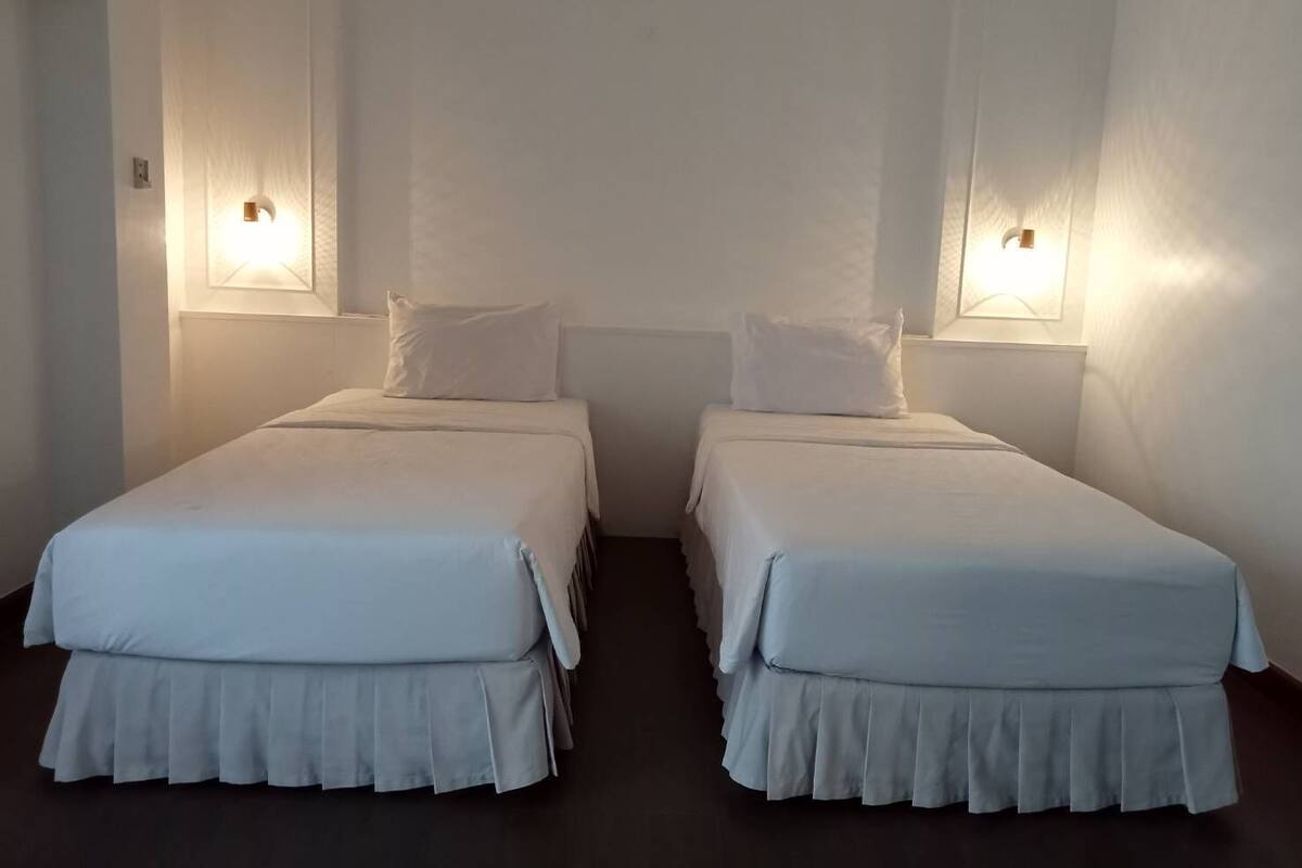 Zwei Einzelbetten mit weißer Bettwäsche und einem dunklen Boden, jeweils mit einer Wandleuchte.