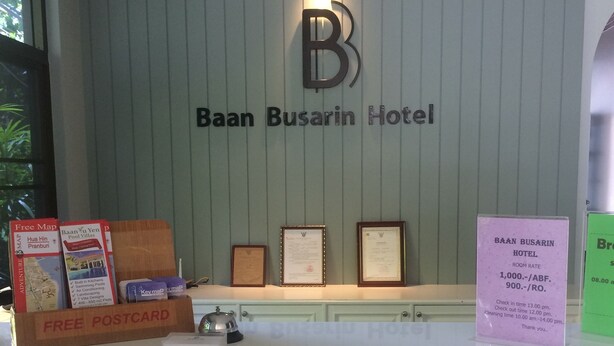 Mostrador de recepción del Hotel Baan Busarin con un cartel que muestra el nombre del hotel, un certificado enmarcado y una caja de 'Postales Gratis'.