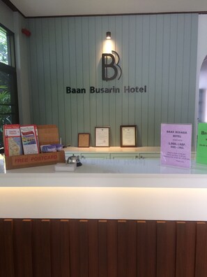 A reception desk at Baan Busarin Hotel with a sign displaying the hotel's name, a framed certificate, and a 'Free Postcard' box. - Baan Busarin Hotel (Hua Hin)