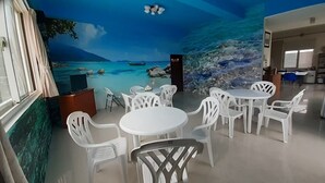 Ein Zimmer mit Wandgemälde einer Strandansicht, weißen Plastikstühlen und Tischen für das Essen sowie einem Deckenventilator.