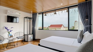 Una habitación de hotel moderna con dos camas, un televisor, aire acondicionado, una mesita con sillas y vistas a edificios a través de ventanas amplias.