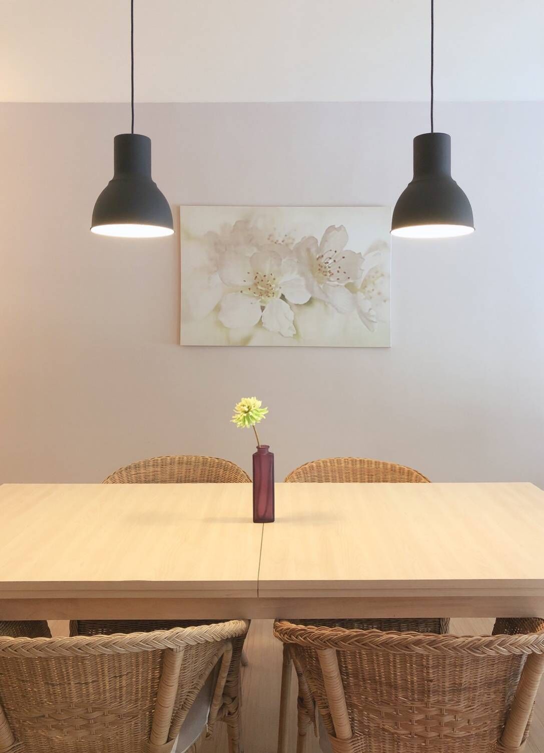 Un coin repas avec une table blanche, des chaises en osier, deux lampes suspendues et un vase avec une fleur.