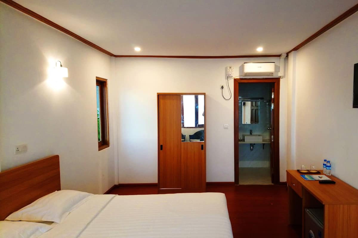 A hotel room with a bed, a desk with a television, a bathroom with a mirror, and an air conditioning unit.