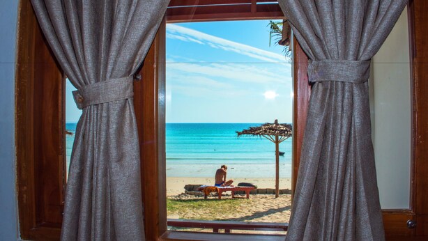 A beach view through a window with curtains, a wooden ceiling, and a person sitting on a beach chair. - Ngapali Paradise Hotel (Thandwe)