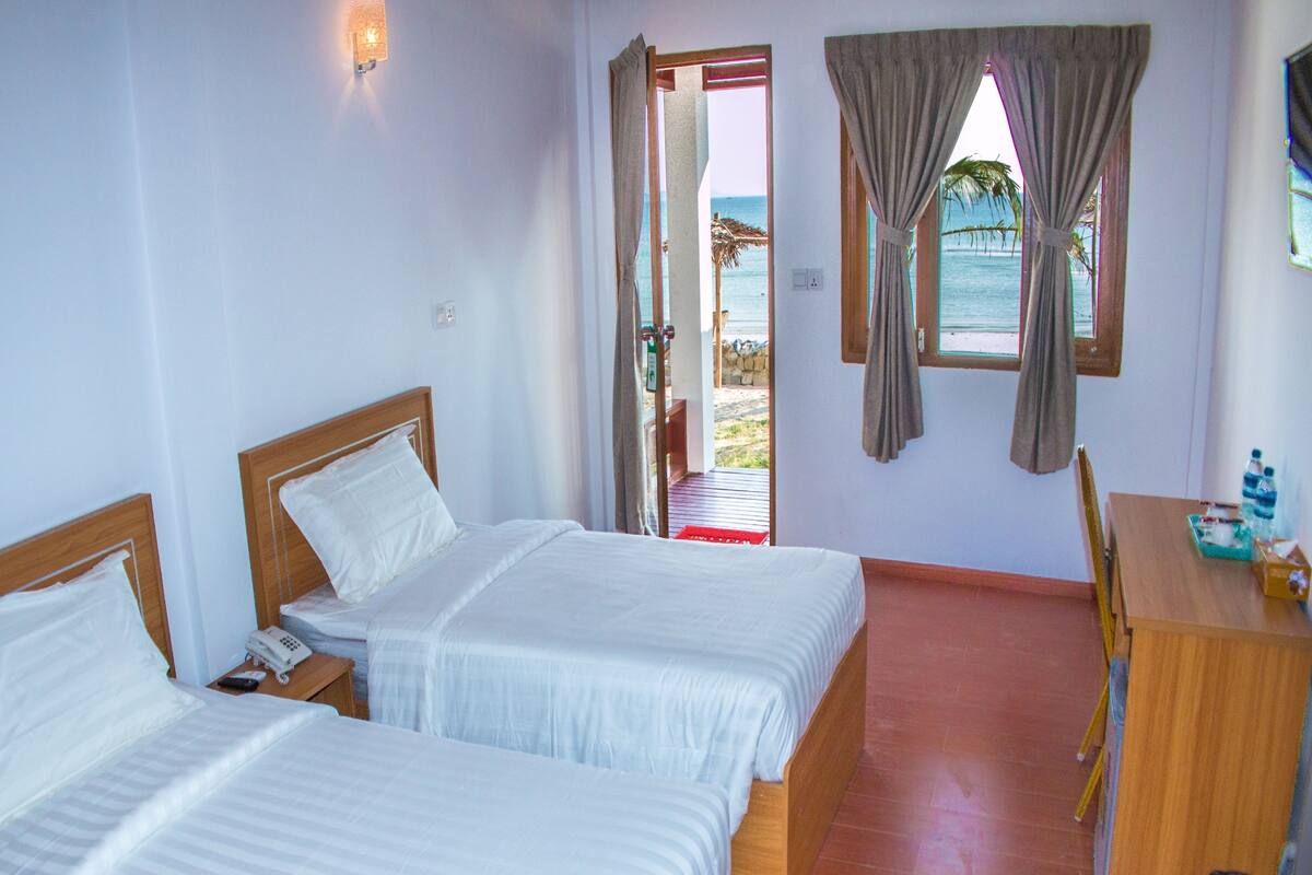 A hotel room with two beds, a wooden desk, and a view of the beach through the window.