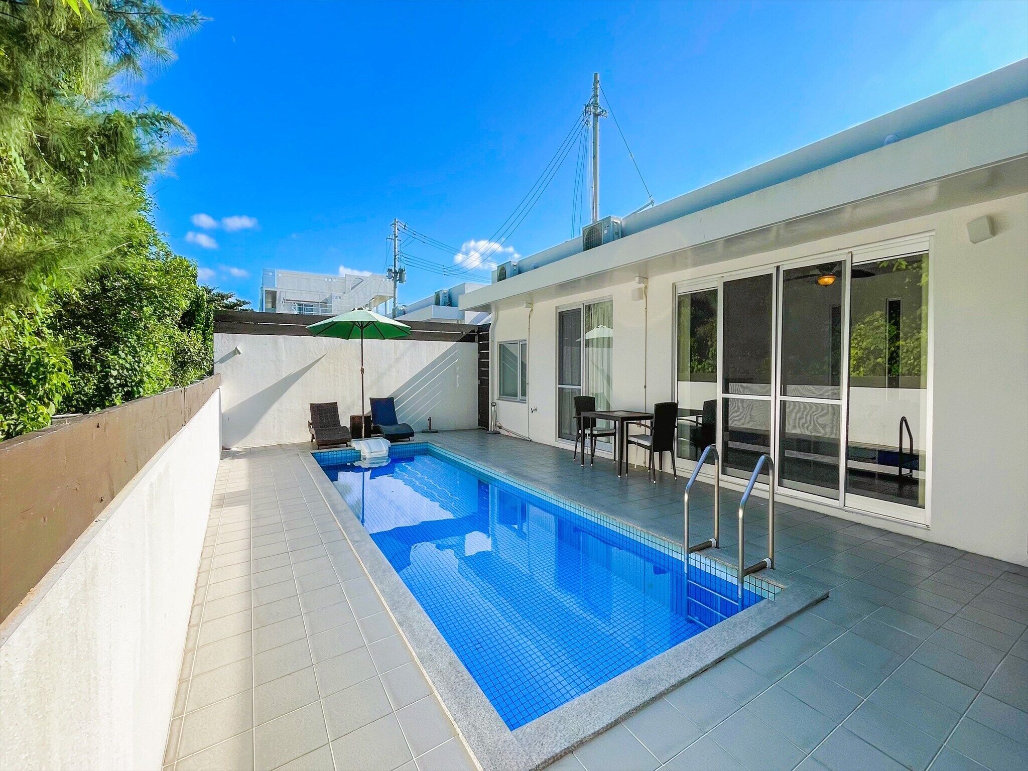 A swimming pool with a tiled deck, a lounge area with chairs and a table, and a view of the sky and buildings in the background.